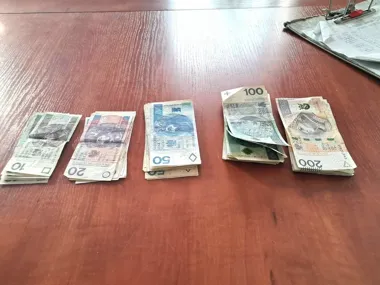 Na drewnianym stole leży pięć stosików polskich banknotów o nominałach 10, 20, 50, 100 i 200 złotych obok metalowej teczki z dokumentami.