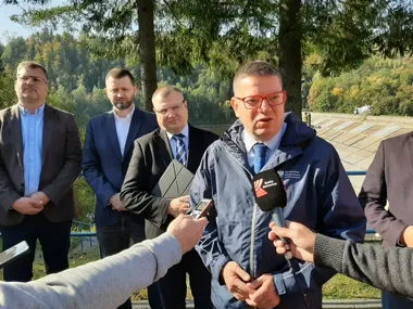 Mężczyzna w kurtce z napisem Ministerstwo Infrastruktury udziela wywiadu dziennikarzom z mikrofonem Radia Katowice na tle zapory wodnej i lasu.